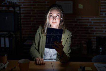 Téléchargez les photos : Blonde femme caucasienne travaillant au bureau la nuit souriant regardant vers le côté et regardant loin en pensant. - en image libre de droit