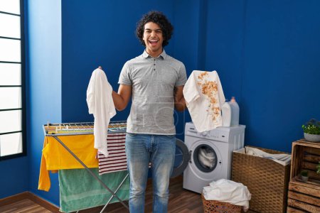 Photo for Hispanic man with curly hair holding clean white t shirt and t shirt with dirty stain smiling and laughing hard out loud because funny crazy joke. - Royalty Free Image
