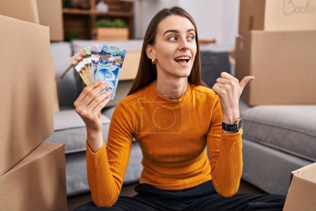 Photo for Young caucasian woman sitting on the floor at new home holding canadian money pointing thumb up to the side smiling happy with open mouth - Royalty Free Image