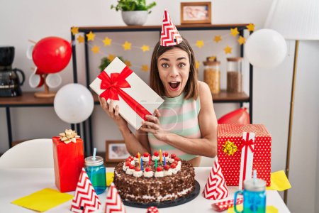 Téléchargez les photos : Femme hispanique célébrant anniversaire avec gâteau tenant cadeau effrayé et choqué avec surprise et expression étonnante, peur et visage excité. - en image libre de droit