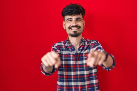 Photo for Young hispanic man with beard standing over red background pointing to you and the camera with fingers, smiling positive and cheerful - Royalty Free Image