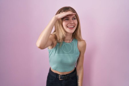Photo for Blonde caucasian woman standing over pink background very happy and smiling looking far away with hand over head. searching concept. - Royalty Free Image