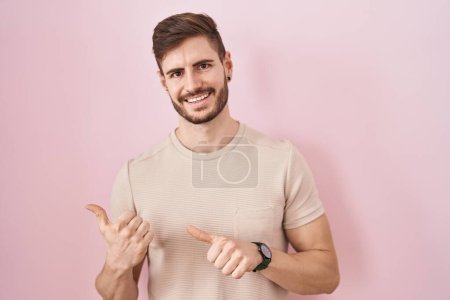 Foto de Hombre hispano con barba de pie sobre fondo rosa apuntando hacia atrás con la mano y los pulgares hacia arriba, sonriendo confiado - Imagen libre de derechos