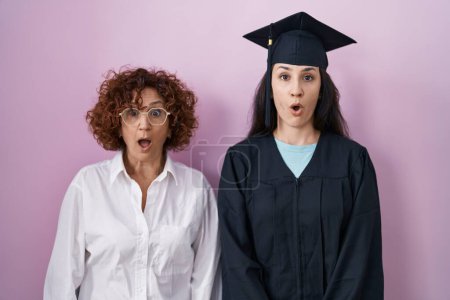 Foto de Madre e hija hispanas con gorra de graduación y bata de ceremonia asustadas y conmocionadas con expresión sorpresa, miedo y cara emocionada. - Imagen libre de derechos