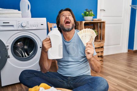 Foto de Guapo hombre de mediana edad ahorrando dinero de ropa detergente enojado y loco gritando frustrado y furioso, gritando con ira mirando hacia arriba. - Imagen libre de derechos