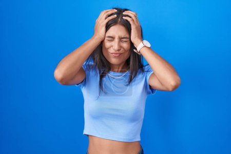 Photo for Brunette young woman standing over blue background suffering from headache desperate and stressed because pain and migraine. hands on head. - Royalty Free Image