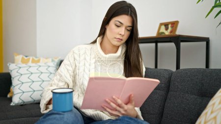 Téléchargez les photos : Jeune belle hispanique femme lecture livre et boire du café assis sur le canapé à la maison - en image libre de droit
