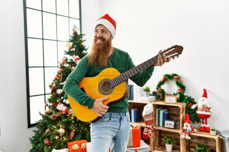 Photo for Young redhead man playing classical guitar standing by christmas tree at home - Royalty Free Image