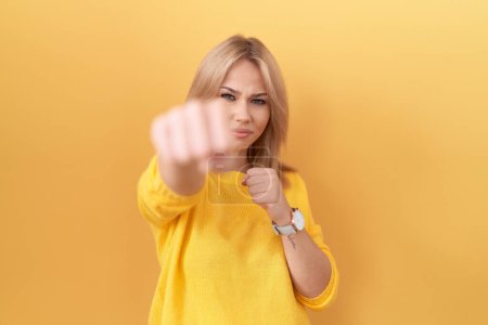 Foto de Mujer caucásica joven con suéter amarillo golpeando puño para luchar, ataque agresivo y enojado, amenaza y violencia - Imagen libre de derechos