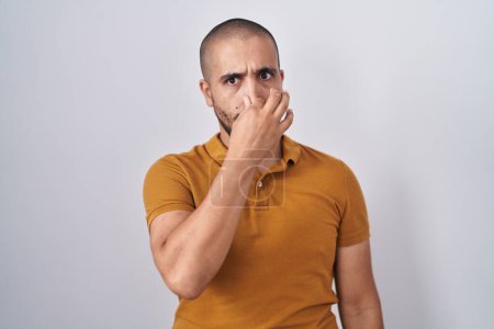 Téléchargez les photos : Homme hispanique avec la barbe debout sur fond blanc sentant quelque chose de puant et dégoûtant, odeur intolérable, retenant souffle avec les doigts sur le nez. mauvaise odeur - en image libre de droit
