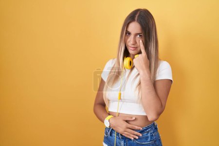 Téléchargez les photos : Jeune femme blonde debout sur fond jaune portant des écouteurs pointant vers l'œil vous regardant geste, expression suspecte - en image libre de droit