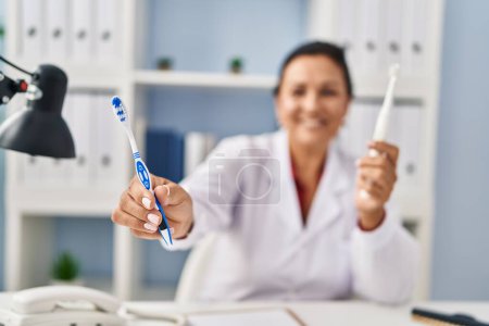 Téléchargez les photos : Middle age hispanic woman dentist holding toothbrushes at clinic - en image libre de droit