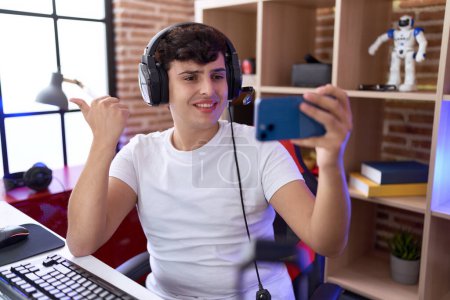 Téléchargez les photos : Jeune homme non binaire jouant à des jeux vidéo avec smartphone pointant le pouce vers le côté souriant heureux avec la bouche ouverte - en image libre de droit