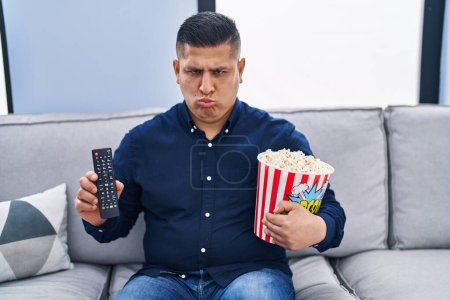 Photo for Hispanic young man eating popcorn using tv control depressed and worry for distress, crying angry and afraid. sad expression. - Royalty Free Image