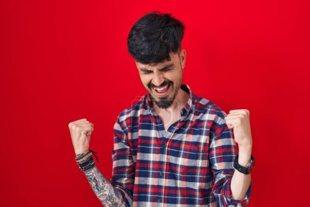Photo for Young hispanic man with beard standing over red background celebrating surprised and amazed for success with arms raised and eyes closed. winner concept. - Royalty Free Image