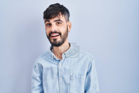 Téléchargez les photos : Jeune homme hispanique avec la barbe debout sur fond bleu souriant gai avec les bras ouverts comme accueil amical, salutations positives et confiantes - en image libre de droit