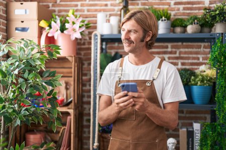 Photo for Young blond man florist smiling confident using smartphone at flower shop - Royalty Free Image