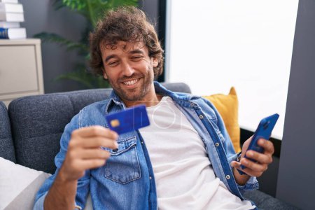 Photo for Young hispanic man using smartphone and credit card sitting on sofa at home - Royalty Free Image