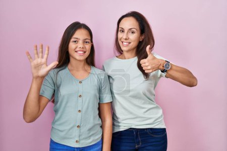 Foto de Joven madre e hija de pie sobre fondo rosa mostrando y señalando hacia arriba con los dedos número seis mientras sonríe confiado y feliz. - Imagen libre de derechos