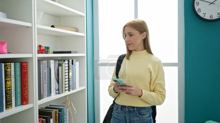 Téléchargez les photos : Jeune étudiante blonde à la recherche d'un livre en utilisant un smartphone dans une salle de classe universitaire - en image libre de droit