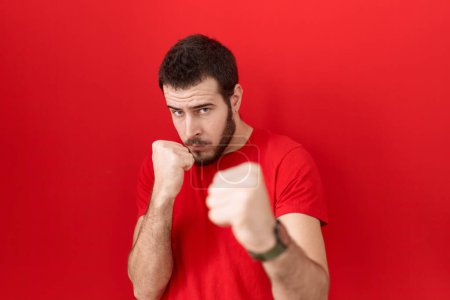 Foto de Joven hombre hispano con camiseta roja casual listo para luchar con el gesto de defensa del puño, cara enojada y molesta, miedo al problema - Imagen libre de derechos