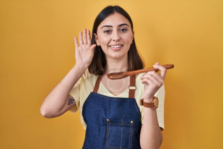 Foto de Chica hispana comiendo cuchara de madera saludable renunciando a decir hola feliz y sonriente, gesto de bienvenida amistoso - Imagen libre de derechos
