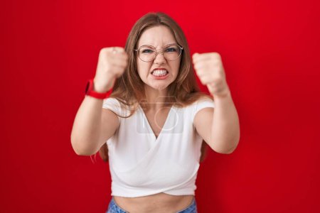 Téléchargez les photos : Jeune femme caucasienne debout sur fond rouge en colère et fou levant les poings frustrés et furieux tout en criant de colère. rage et concept agressif. - en image libre de droit