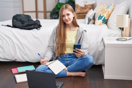 Téléchargez les photos : Young caucasian woman using smartphone and laptop writing on notebook at bedroom - en image libre de droit