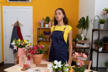 Téléchargez les photos : Jeune afro-américaine fleuriste debout avec une expression sérieuse à la boutique de fleurs - en image libre de droit
