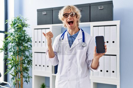 Téléchargez les photos : Jeune homme médecin montrant écran smartphone pointant pouce vers le côté souriant heureux avec bouche ouverte - en image libre de droit