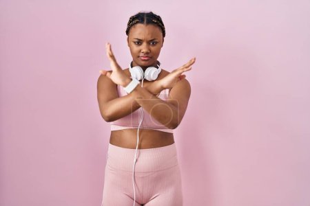 Téléchargez les photos : Femme afro-américaine avec des tresses portant des vêtements de sport et des écouteurs expression de rejet croisant les bras faisant signe négatif, visage en colère - en image libre de droit