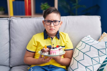Téléchargez les photos : Jeune enfant hispanique tenant bonbons gâteau sceptique et nerveux, fronçant les sourcils bouleversé en raison de problèmes. personne négative. - en image libre de droit