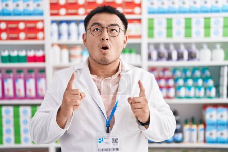 Téléchargez les photos : Jeune homme chinois travaillant à la pharmacie pharmacie étonné et surpris de regarder vers le haut et pointant avec les doigts et les bras levés. - en image libre de droit