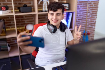 Téléchargez les photos : Non binaire homme streamer sourire confiant faire selfie par smartphone à la salle de jeux - en image libre de droit