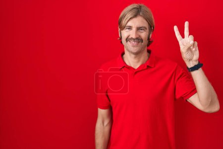 Téléchargez les photos : Homme caucasien avec moustache debout sur fond rouge montrant et pointant vers le haut avec les doigts numéro deux tout en souriant confiant et heureux. - en image libre de droit