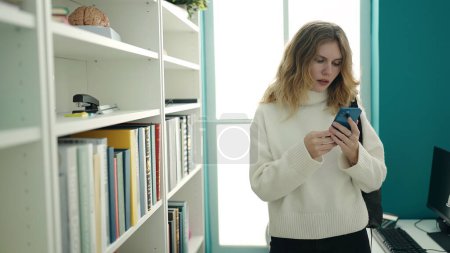 Téléchargez les photos : Jeune étudiante blonde utilisant un smartphone debout à la bibliothèque de l'université - en image libre de droit