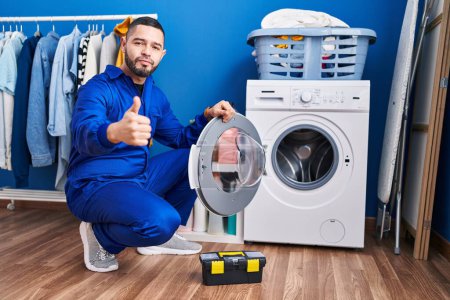 Téléchargez les photos : Réparateur hispanique travaillant sur la machine à laver souriant heureux et positif, pouce levé faire excellent et signe d'approbation - en image libre de droit