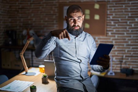 Photo for Young hispanic man with beard and tattoos working at the office at night pointing down looking sad and upset, indicating direction with fingers, unhappy and depressed. - Royalty Free Image