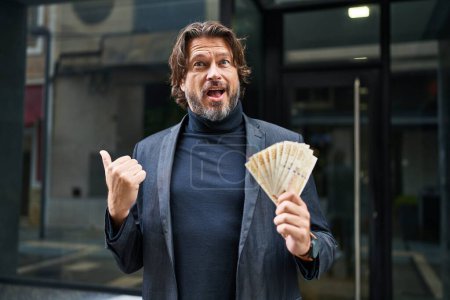 Foto de Hombre guapo de mediana edad sosteniendo billetes de 100 coronas danesas apuntando con el pulgar hacia el costado sonriendo feliz con la boca abierta - Imagen libre de derechos