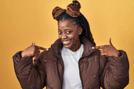 Photo for African woman with braided hair standing over yellow background looking confident with smile on face, pointing oneself with fingers proud and happy. - Royalty Free Image