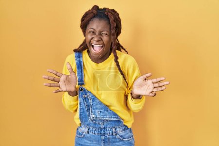 Foto de Mujer africana de pie sobre fondo amarillo celebrando loco y sorprendido por el éxito con los brazos levantados y los ojos abiertos gritando emocionado. concepto ganador - Imagen libre de derechos