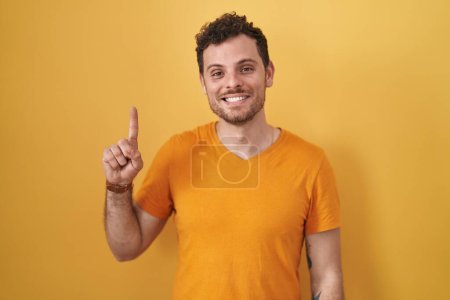 Foto de Joven hombre hispano de pie sobre fondo amarillo mostrando y señalando con el dedo número uno mientras sonríe confiado y feliz. - Imagen libre de derechos