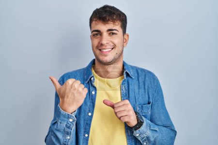 Téléchargez les photos : Jeune homme hispanique debout sur fond bleu pointant vers le dos derrière avec la main et les pouces levés, souriant confiant - en image libre de droit