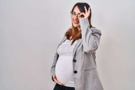 Téléchargez les photos : Femme d'affaires enceinte debout sur fond blanc souriant heureux faisant signe ok avec la main sur les yeux regardant à travers les doigts - en image libre de droit