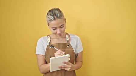Foto de Mujer hispana hermosa joven con delantal escrito en cuaderno sobre fondo amarillo aislado - Imagen libre de derechos