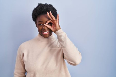 Foto de Mujer afroamericana de pie sobre fondo azul haciendo buen gesto con la mano sonriendo, los ojos mirando a través de los dedos con la cara feliz. - Imagen libre de derechos