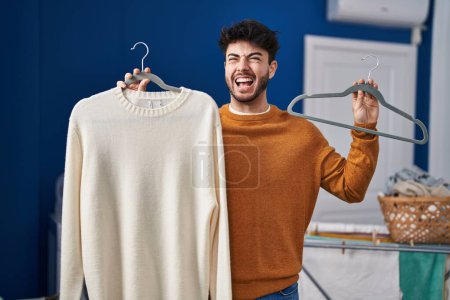 Photo for Hispanic man with beard holding sweater on hanger at laundry room angry and mad screaming frustrated and furious, shouting with anger. rage and aggressive concept. - Royalty Free Image