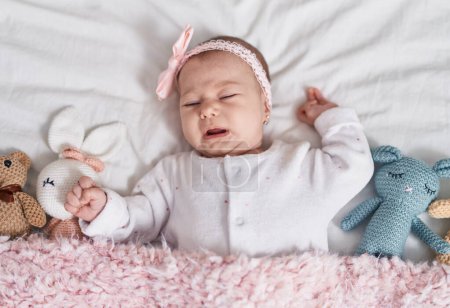 Foto de Adorable bebé caucásico acostado en la cama con muñecas llorando en el dormitorio - Imagen libre de derechos