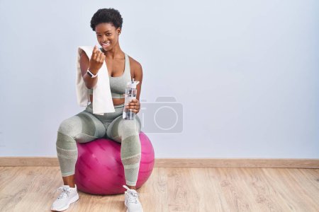 Téléchargez les photos : Femme afro-américaine vêtue de vêtements de sport assis sur pilates ball beckoning venir ici geste avec la main invitant accueillant heureux et souriant - en image libre de droit