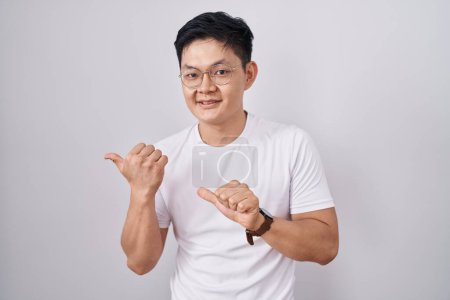 Foto de Joven asiático hombre de pie sobre blanco fondo apuntando a la espalda detrás con la mano y los pulgares hacia arriba, sonriendo confiado - Imagen libre de derechos